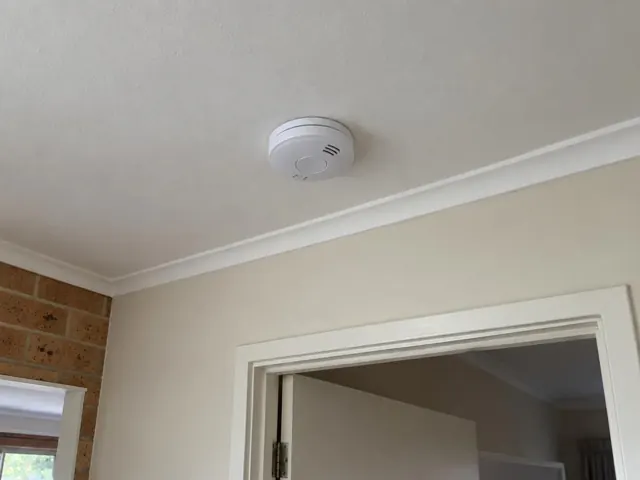 Ceiling-mounted smoke alarm above a doorway in a beige home interior in Brookvale